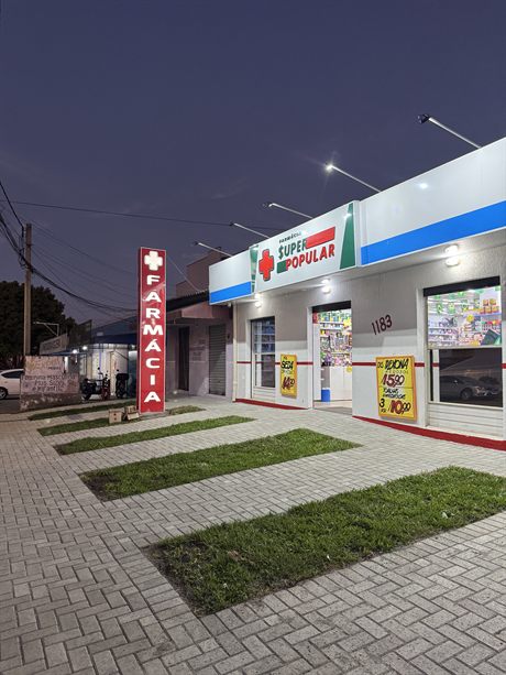 Fachada da Farmácia Super Popular Lindóia à noite, com o letreiro aceso e o totem vermelho escrito Farmácia na calçada.